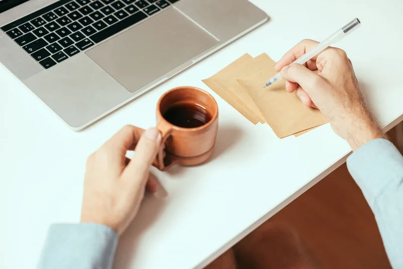 A person taking notes by hand next to a laptop and a cup of coffee