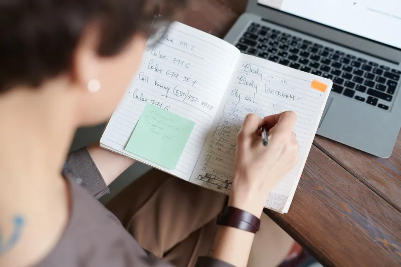 Person writing in a notebook with a laptop on a table, researching their target market